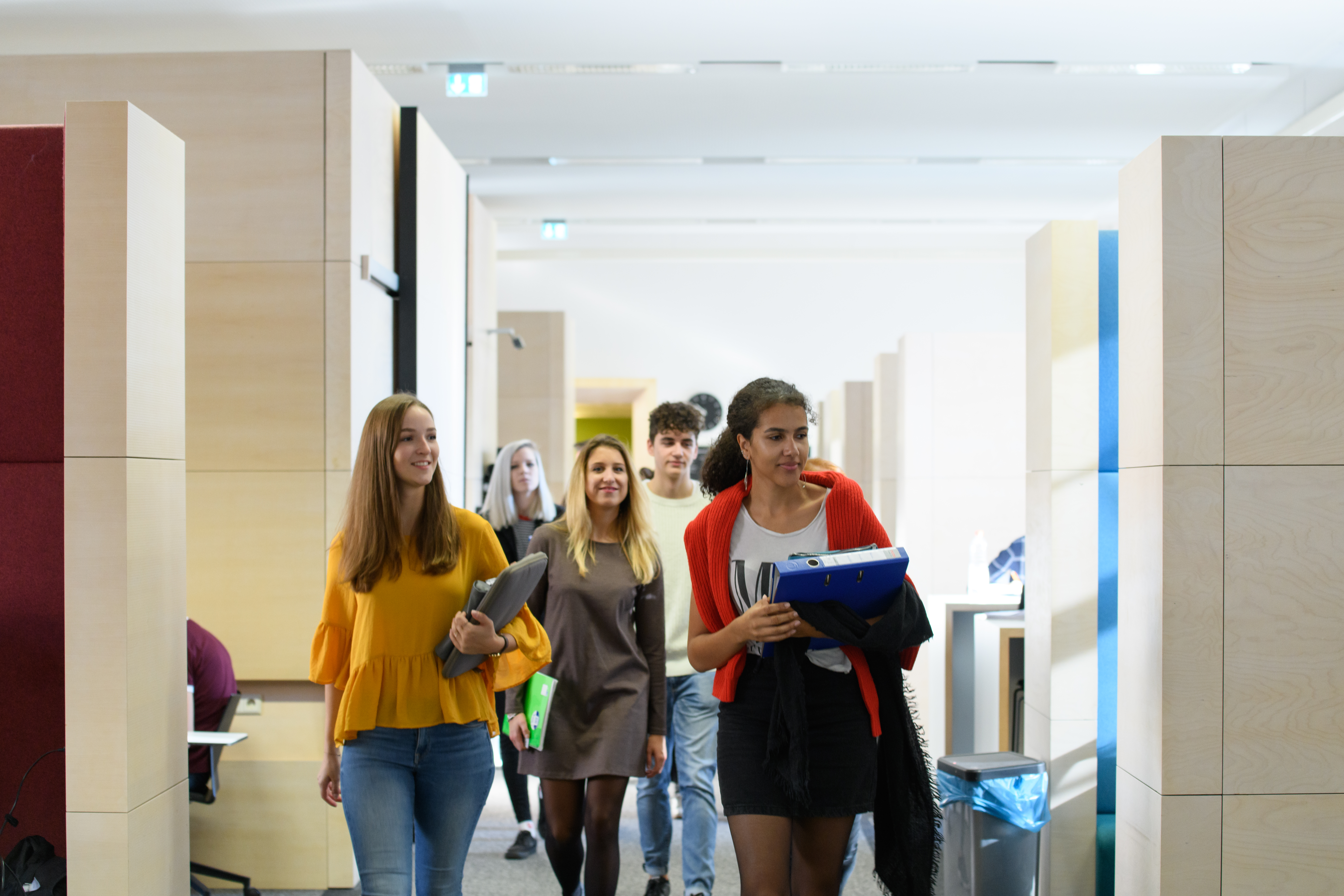 Eine Gruppe Studierender läuft durch das Learning Center im Bibliotheksbereich Schloss Schneckenhof. Sie haben Laptop, Ordner, Collegeblock und Stiftemäppchen dabei.