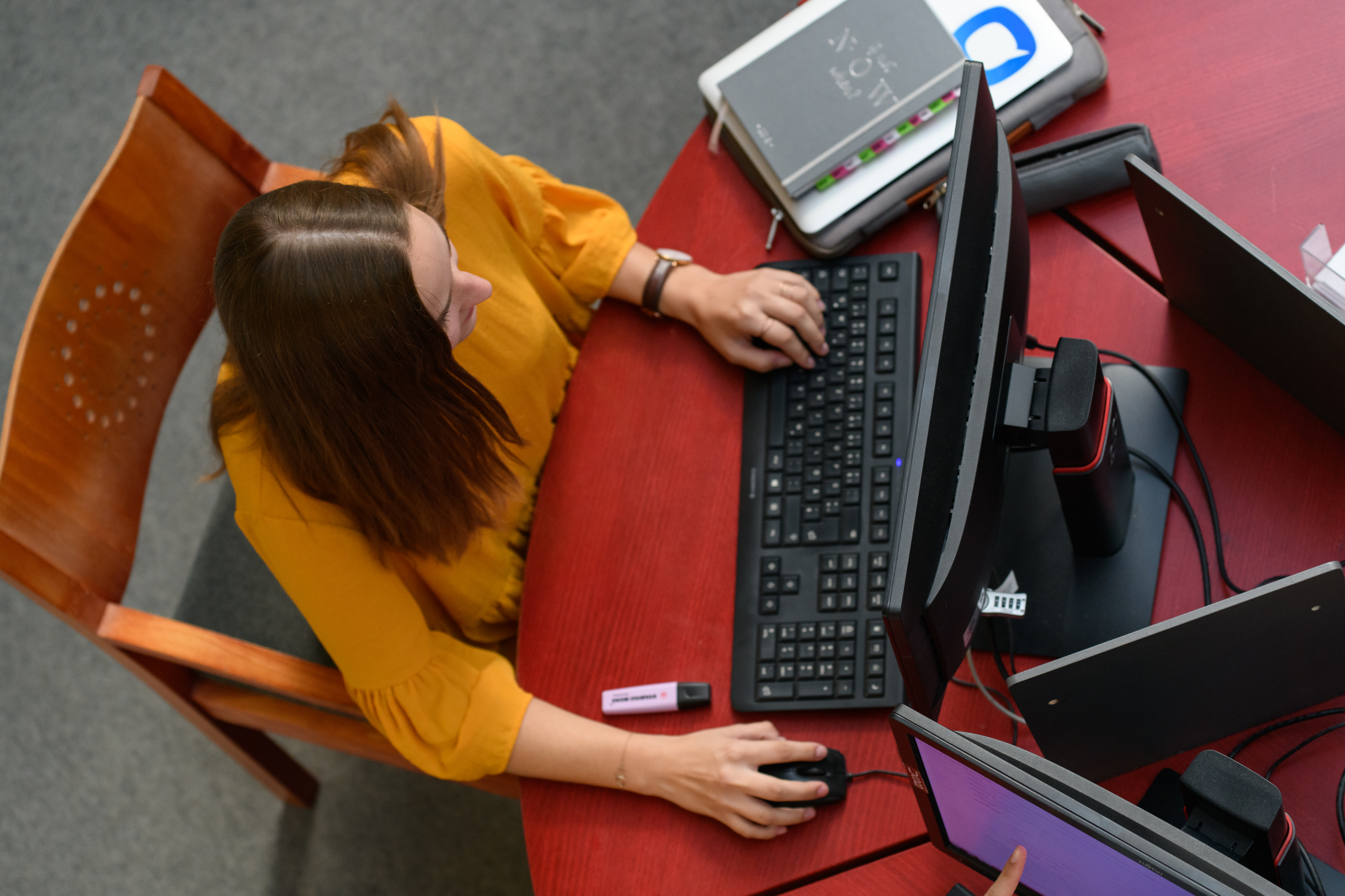 Blick von oben: Eine Studentin sitzt vor einem Recherche-PC an einem Arbeitsplatz im Bibliotheksbereich A3. Ihre linke Hand ist auf der Tastatur, die rechte Hand an der Computermaus. Neben der Tastatur liegt ein Stapel aus Laptophülle, Laptop und Notizbuch, daneben ein Stiftemäppchen. Vor der Tastatur liegt ein pinkfarbener Textmarker. 