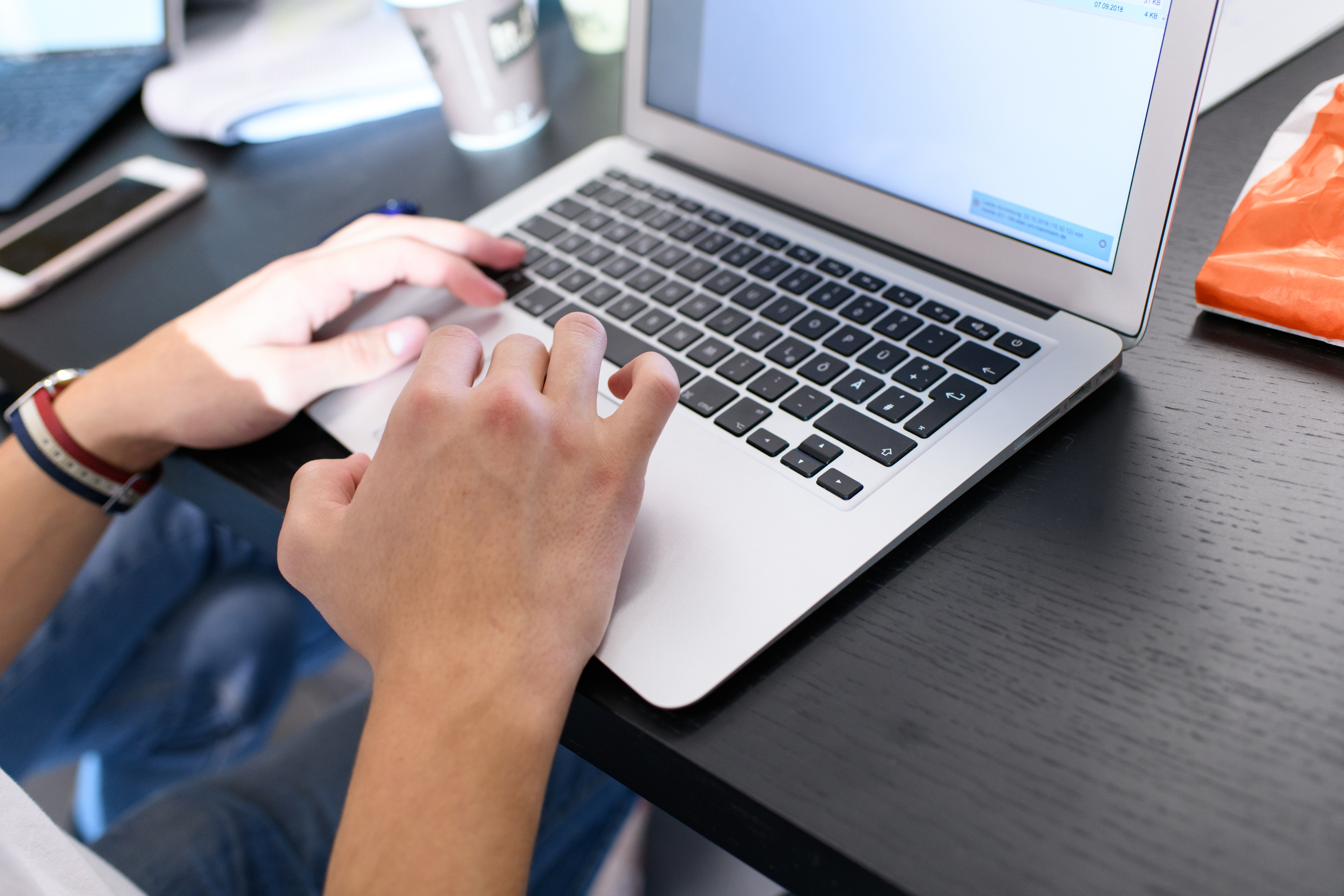 Eine Person, deren Hände im Bild sind, schreibt an einem Laptop. Neben dem Laptop sind ein Smartphone und ein Kaffeebecher auf dem Tisch.