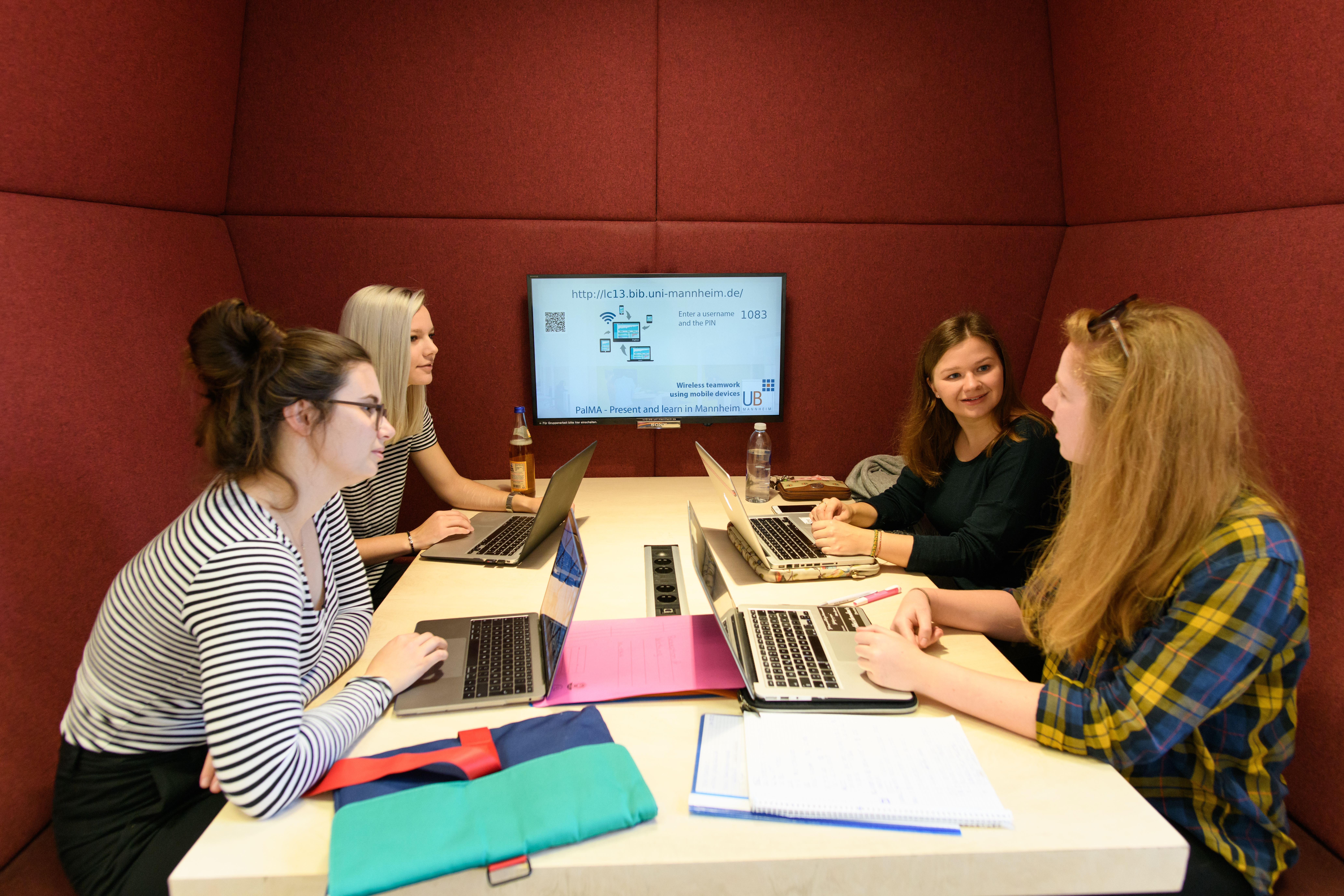 Vier Studierende sitzen in einem roten Kubus im Learning Center im Bibliotheksbereich Schloss Schneckenhof. Sie sind miteinander im Gespräch, jede Person hat ein Laptop vor sich, auf dem Tisch liegen Collegeblöcke und Mappen.