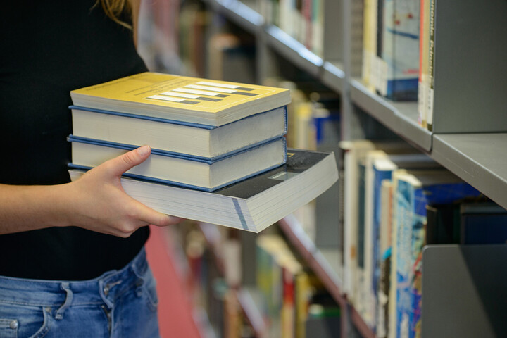 Eine Person steht vor einem Bücherregal und hält einen Stapel aus vier Bücher in den Händen.