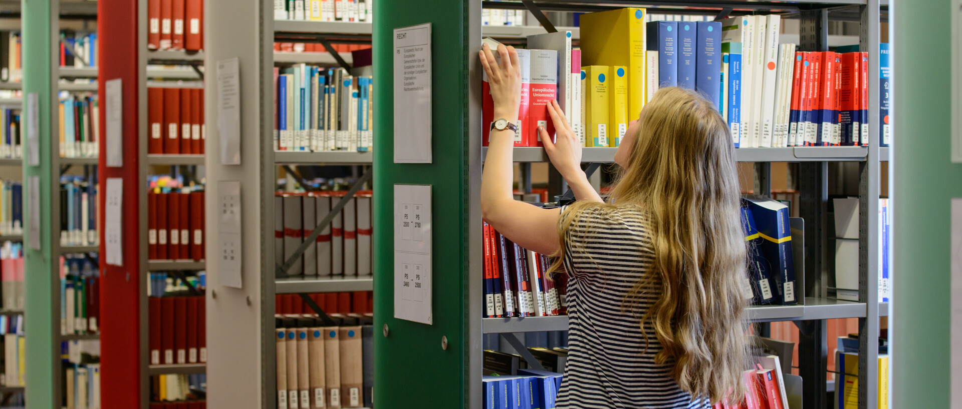Eine Studentin steht vor einem Bücherregal im Bibliotheksbereich Schloss Ehrenhof. Sie holt ein Buch aus dem Regal bzw. stellt es zurück. Dahinter sind weitere Bücherregale mit Regalblenden in Grün-, Rot- und Weißtönen.