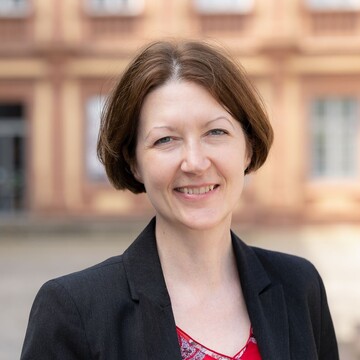 Annette Klein hat halblanges braunes Haar, sie trägt ein rotes Oberteil mit floralem Muster und ein schwarzes Jackett.