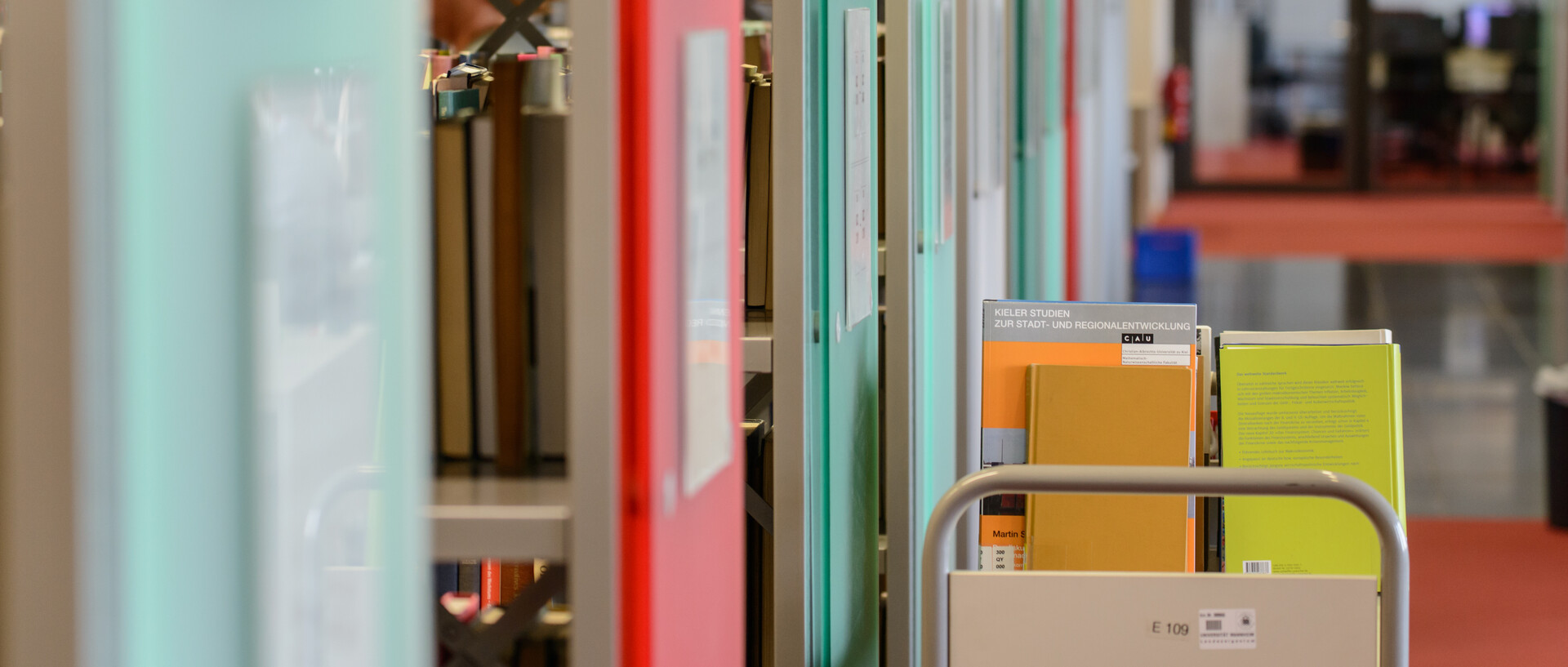 Ein Bücherwagen mit Büchern steht vor Bücherregalen mit grünen und roten Regalfronten im Bibliotheksbereich Schloss Ehrenhof.