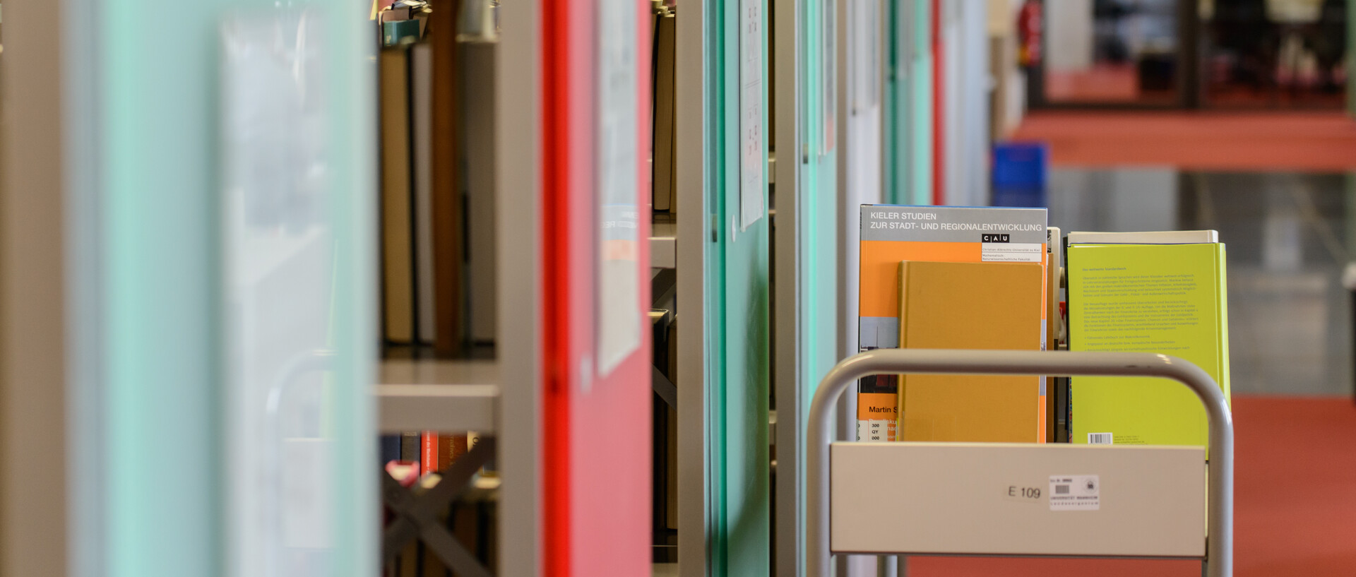 Ein Bücherwagen mit Büchern steht vor Bücherregalen mit grünen und roten Regalfronten im Bibliotheksbereich Schloss Ehrenhof.