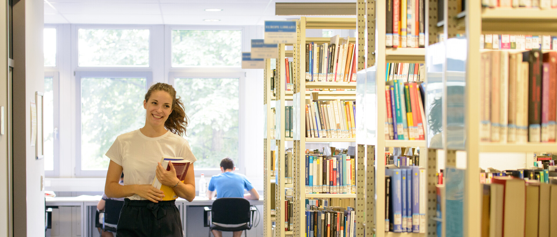 Eine Studentin läuft an Bücherregalen im Bibliotheksbereich A5 entlang. In der linken Armbeuge hält sie ein Buch. Im Hintergrund sitzt ein Student an einem Arbeitsplatz.