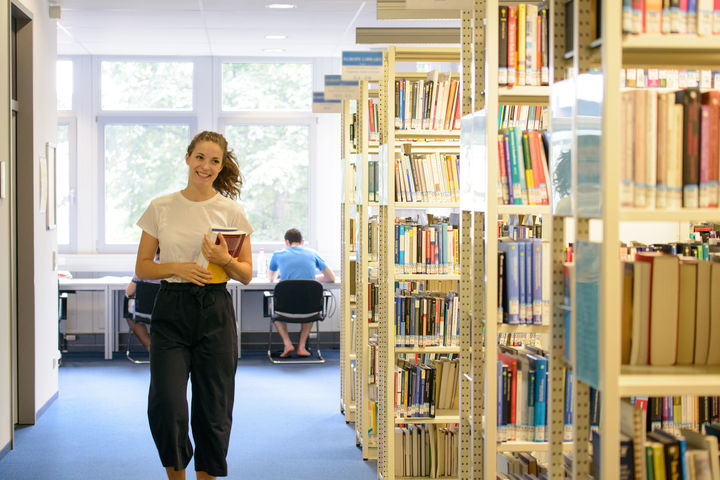 Eine Studentin läuft an Bücherregalen im Bibliotheksbereich A5 entlang. In der linken Armbeuge hält sie ein Buch. Im Hintergrund sitzt ein Student an einem Arbeitsplatz.