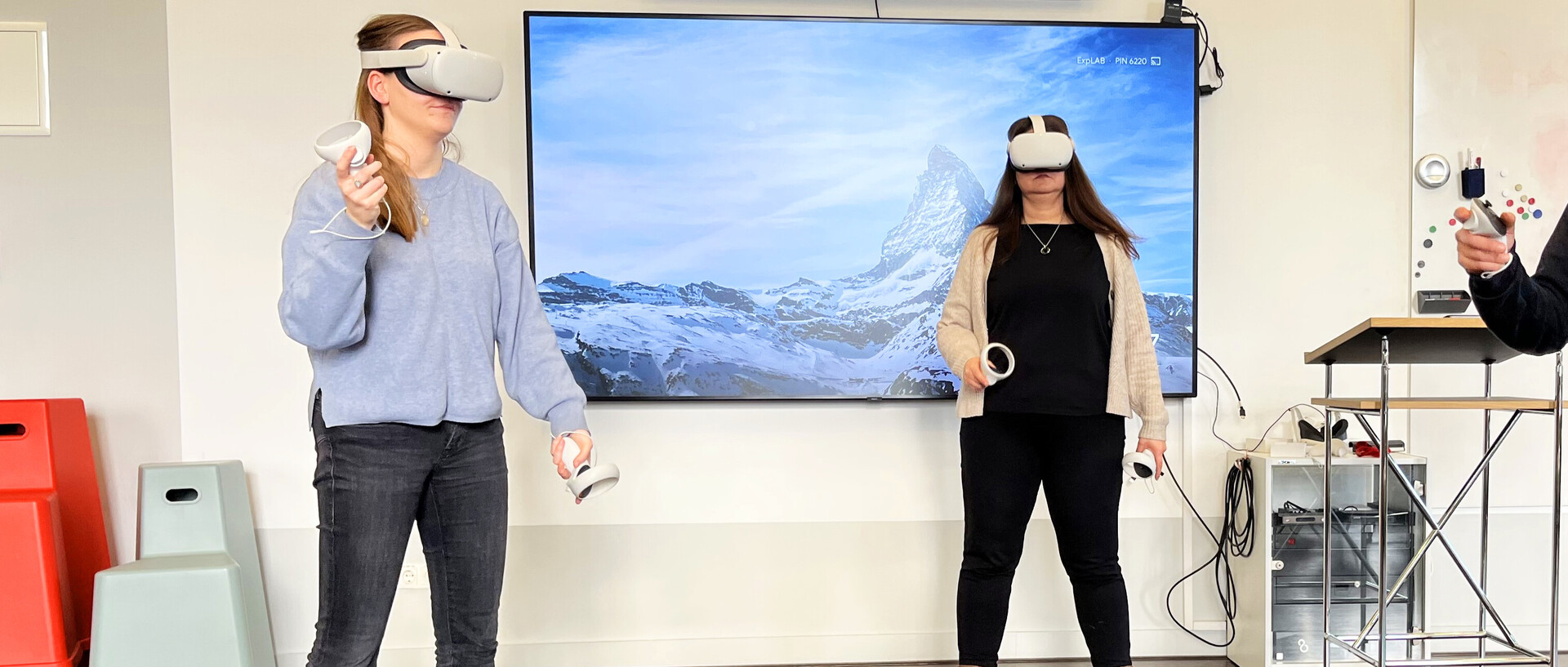 Drei Personen benutzen VR-Brillen im ExpLAB im Bibliotheksbereich Schloss Schneckenhof. An der Wand hängt ein großer Präsentationsmonitor, auf dem eine Schneelandschaft zu erkennen ist.