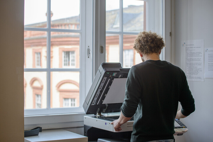 Ein Student benutzt einen Kopierer in der Bibliothek. Die Geräteklappe ist geöffnet. Durch ein großes Sprossenfenster ist im Hintergrund das Schloss zu sehen. 