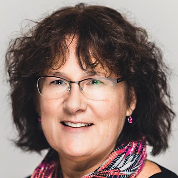 Barbara Dreisiebner hat kinnlanges braunes Haar, sie trägt eine Brille, Ohrringe, ein Halstuch mit Muster in Pink, Rot, Schwarz und Grau und einen schwarzen Pullover.