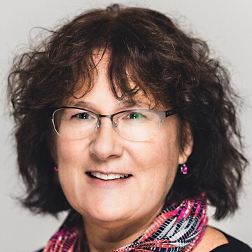 Barbara Dreisiebner hat kinnlanges braunes Haar, sie trägt eine Brille, Ohrringe, ein Halstuch mit Muster in Pink, Rot, Schwarz und Grau und einen schwarzen Pullover. 