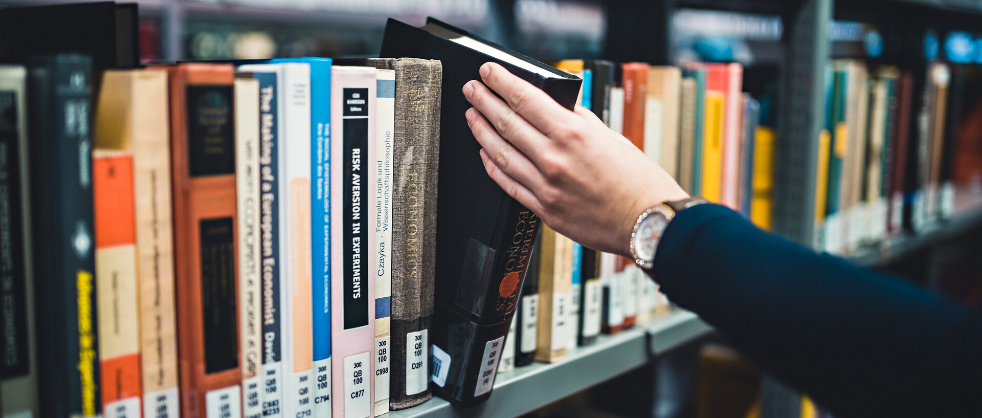 Eine Person, deren linke Hand und linker Arm im Bild sind, nimmt ein Buch aus einem Regal in der Bibliothek bzw. stellt es zurück. Die Person trägt eine Armbanduhr und einen dunkelblauen Pullover.