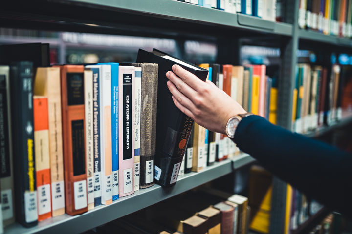 Eine Person, deren linke Hand und linker Arm im Bild sind, nimmt ein Buch aus einem Regal in der Bibliothek bzw. stellt es zurück. Die Person trägt eine Armbanduhr und einen dunkelblauen Pullover.