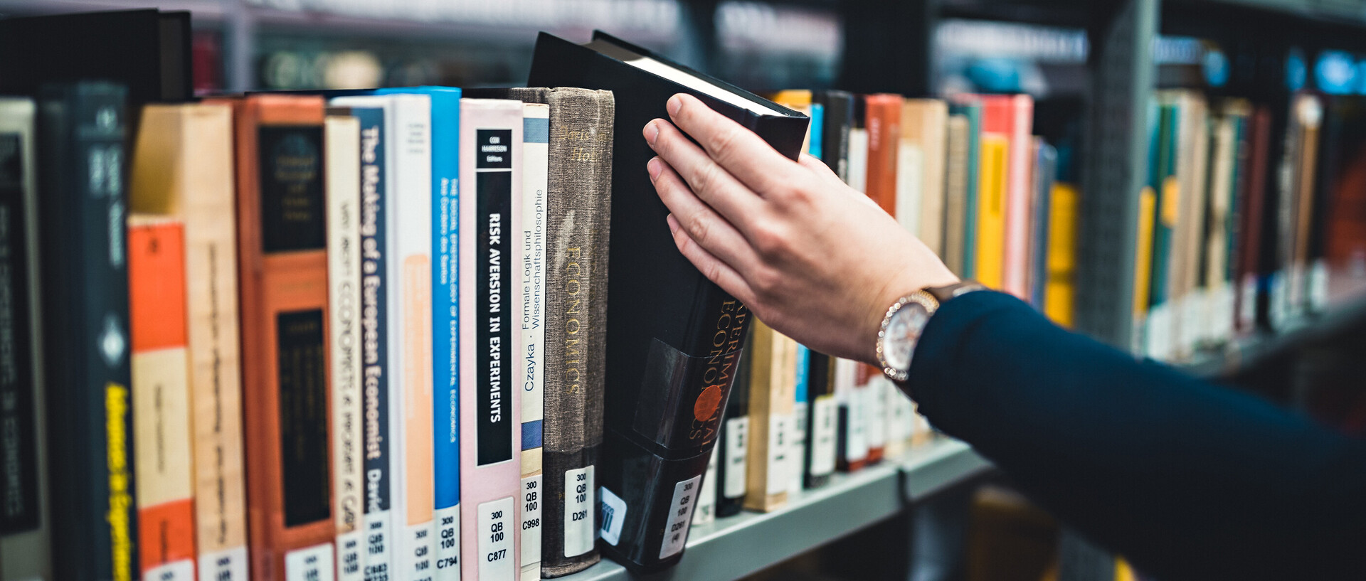 Eine Person, deren rechter Arm und rechte Hand im Bild sind, nimmt ein Buch aus dem Bücherregal in der Bibliothek bzw. stellt es zurück. Die Person trägt eine Armbanduhr und einen dunkelblauen Pullover.
