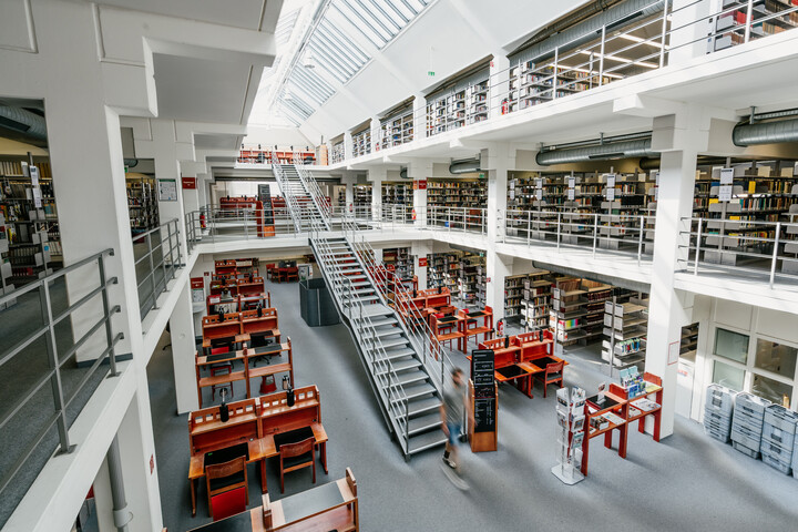 Bibliotheksbereich A3 mit vielen Arbeitsplätzen und Bücherregalreihen im Erdgeschoss, dem ersten und zweiten Stock mit Bücherregalreihen, einer langen Treppe vom Erdgeschoss bis in den zweiten Stock und einem sonnigen Glasdach.