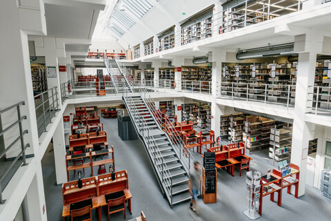 Bibliotheksbereich A3 mit vielen Arbeitsplätzen und Bücherregalreihen im Erdgeschoss, dem ersten und zweiten Stock mit Bücherregalreihen, einer langen Treppe vom Erdgeschoss bis in den zweiten Stock und einem sonnigen Glasdach.