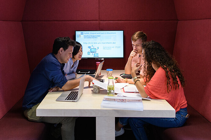 Vier Studierende sitzen in einem roten Kubus im Learning Center im Bibliotheksbereich Schloss Schneckenhof. An der Rückwand des Kubus hängt ein PalMA-Team-Monitor.
