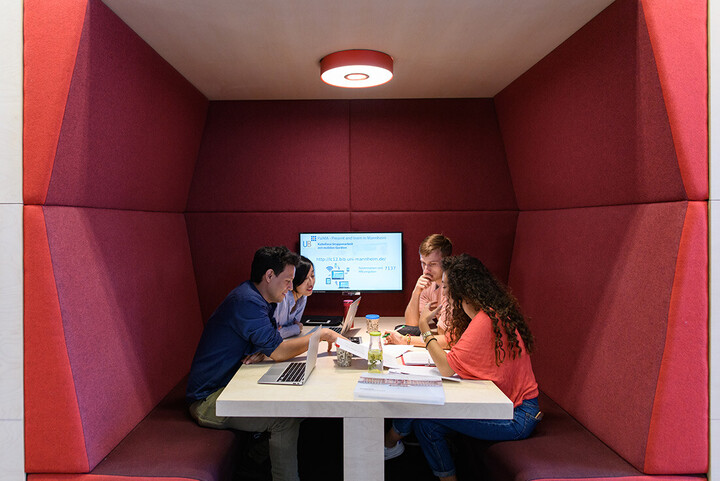Vier Studierende sitzen in einem roten Kubus im Learning Center im Bibliotheksbereich Schloss Schneckenhof. An der Rückwand des Kubus hängt ein PalMA-Team-Monitor.
