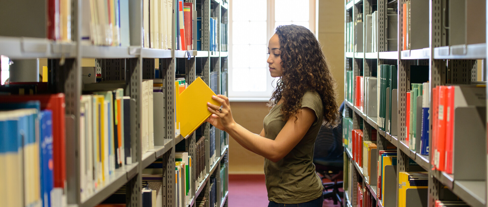 Eine Studentin steht zwischen zwei Bücherregalen in der Bibliothek und nimmt ein Buch aus dem Regal bzw. stellt es zurück. Im Hintergrund ist ein großes Fenster.