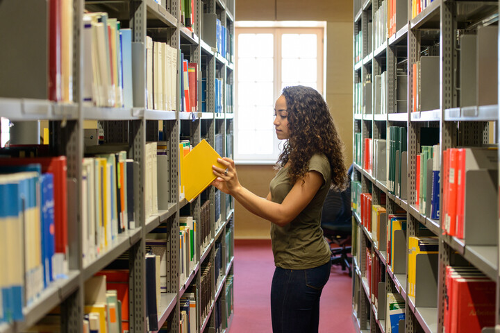 Eine Studentin steht zwischen zwei Bücherregalen in der Bibliothek und nimmt ein Buch aus dem Regal bzw. stellt es zurück. Im Hintergrund ist ein großes Fenster.