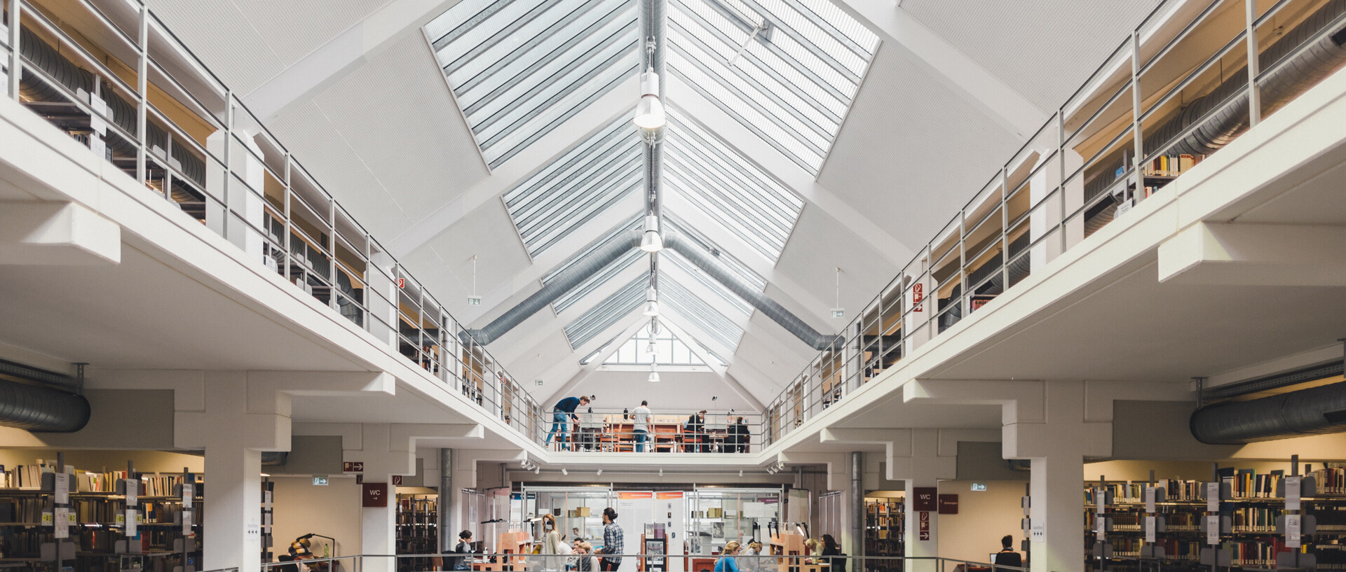 Blick auf die Ausstellungsfläche, die Galerie mit Bücherregalen und das Glasdach im Bibliotheksbereich A3.