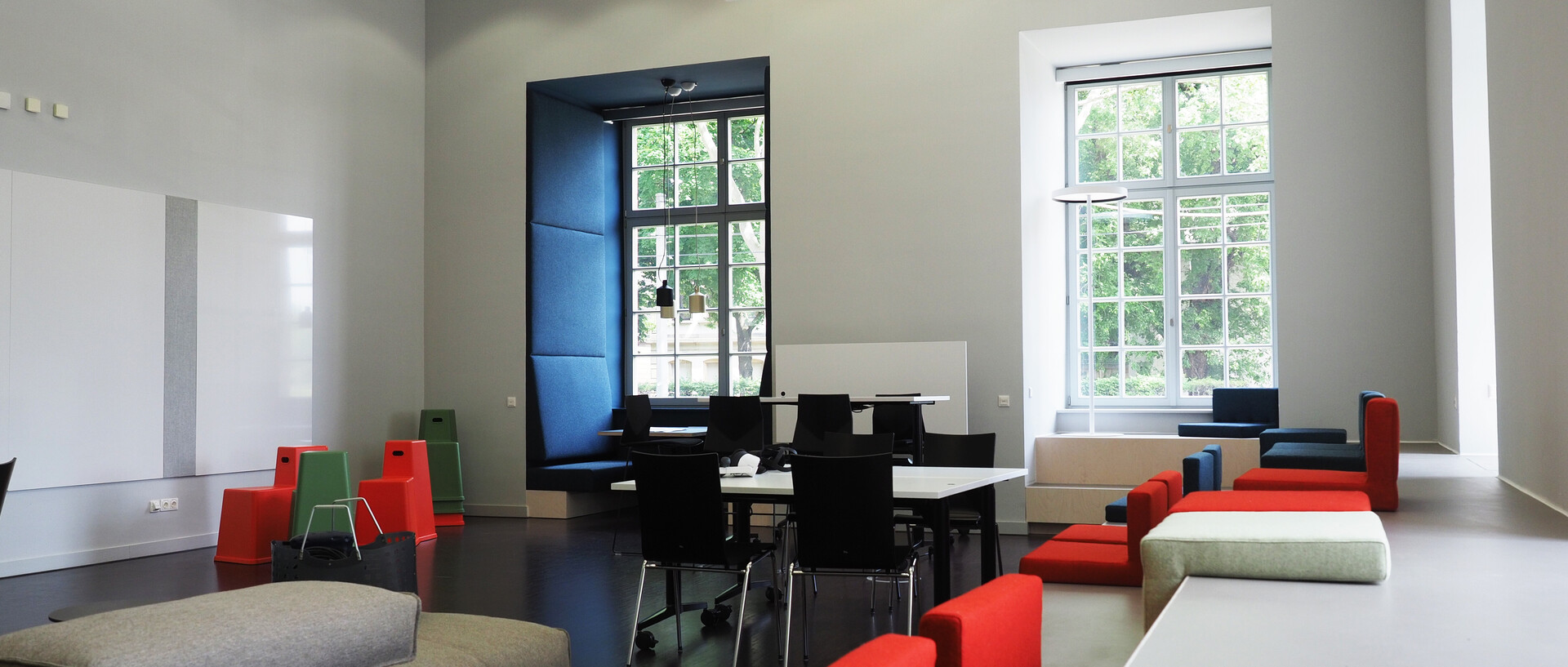 Blick in das ExpLAB im Bibliotheksbereich Schloss Schneckenhof mit Sitzpodesten, großen Fenstern, Tischen, Stühlen, Sofa und Sitzkissen.
