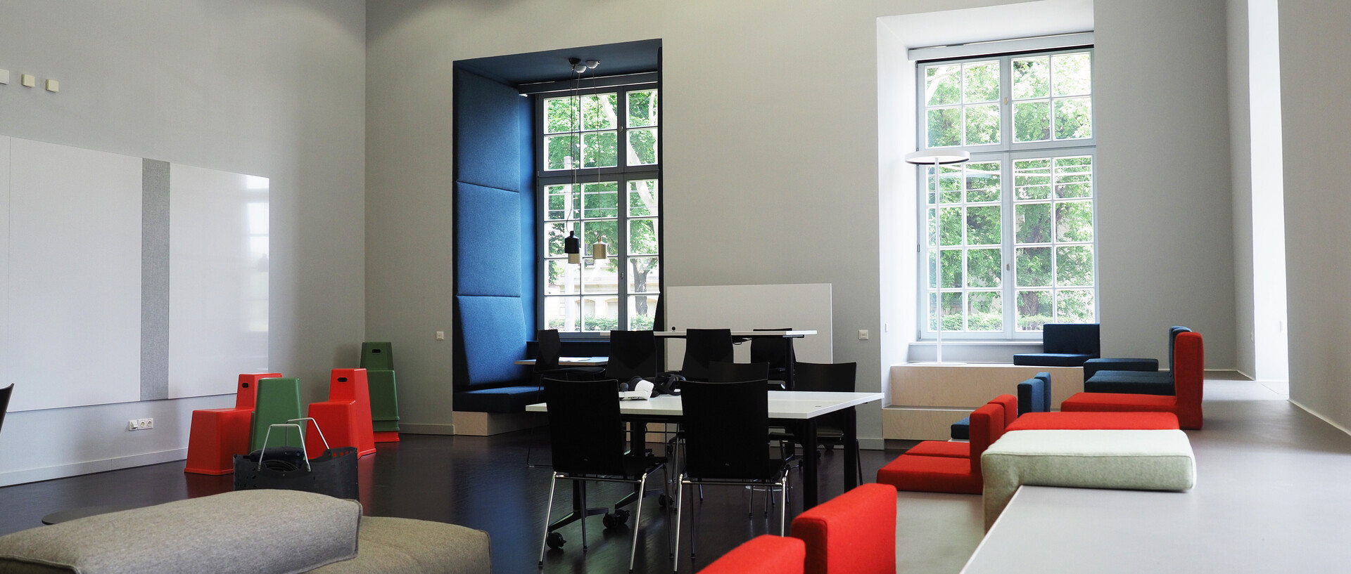 Blick in das ExpLAB im Bibliotheksbereich Schloss Schneckenhof mit Sitzpodesten, großen Fenstern, Tischen, Stühlen, Sofa und Sitzkissen.