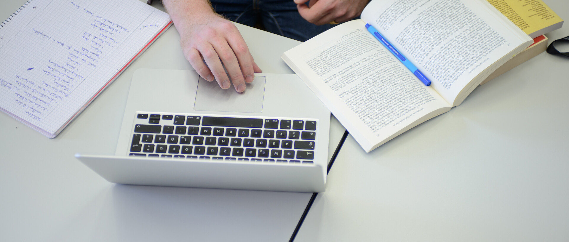 Auf Tischen in einem Seminarraum liegen ein Schreibblock, ein Laptop und Bücher. Am Tisch sitzt eine Person, deren Hände im Bild sind.