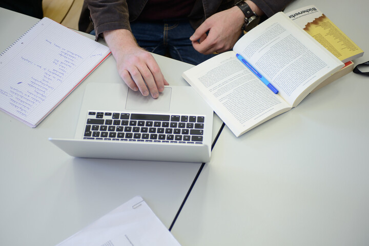Auf Tischen in einem Seminarraum liegen ein Schreibblock, ein Laptop und Bücher. Am Tisch sitzt eine Person, deren Hände im Bild sind.