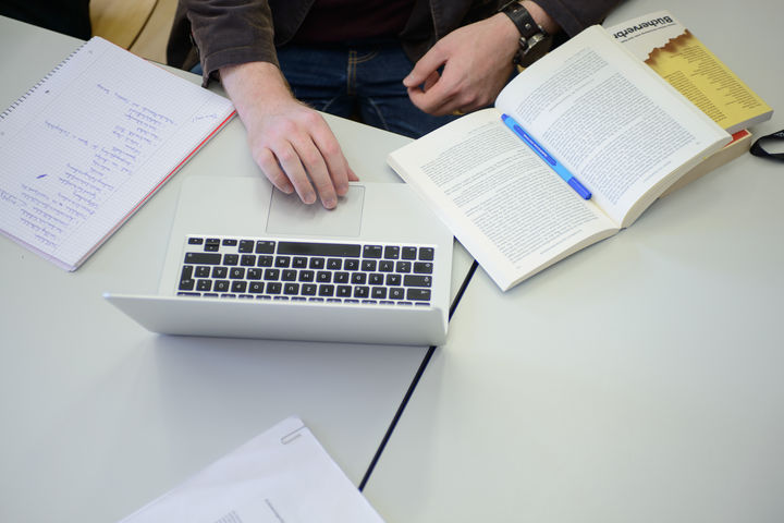 Auf Tischen in einem Seminarraum liegen ein Schreibblock, ein Laptop und Bücher. Am Tisch sitzt eine Person, deren Hände im Bild sind.