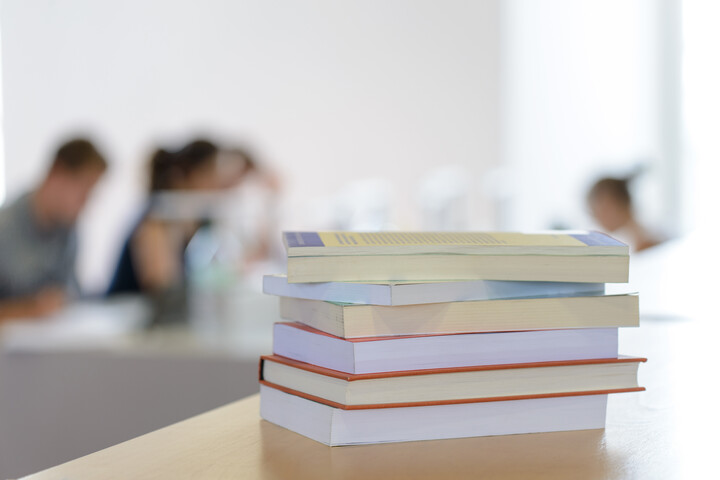 Ein Stapel Bücher liegt auf einem Tisch. Im Hintergrund sind verschwommen Personen an Schreibtischen zu erkennen.
