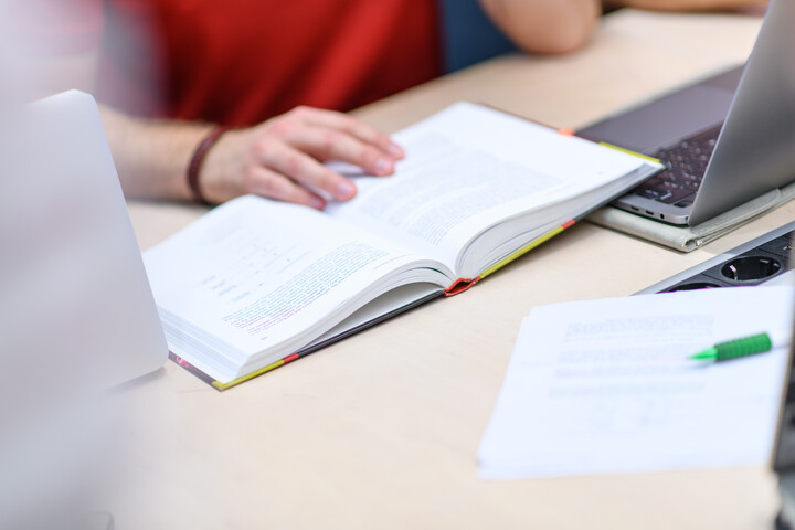 Arbeitsplatz in der Bibliothek, eine Person hat ein aufgeschlagenes Buch und einen Laptop vor sich auf dem Tisch, dahinter liegen Ausdrucke und ein grüner Stift.