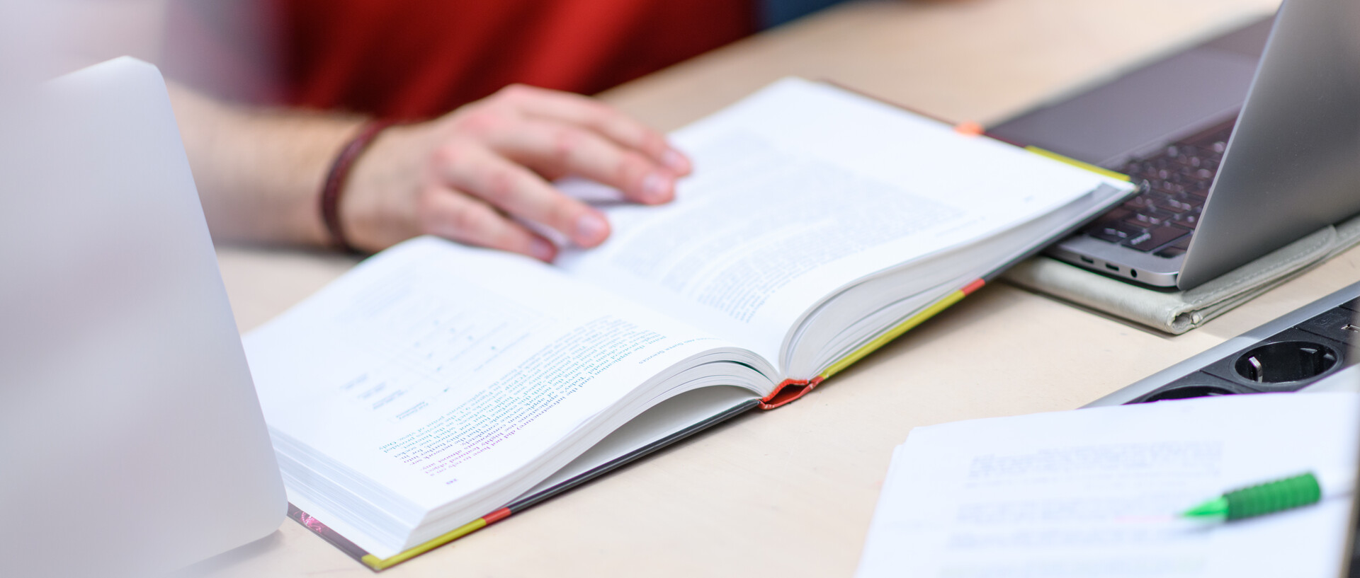Arbeitsplatz in der Bibliothek, eine Person hat ein aufgeschlagenes Buch und einen Laptop vor sich auf dem Tisch, dahinter liegen Ausdrucke und ein grüner Stift.