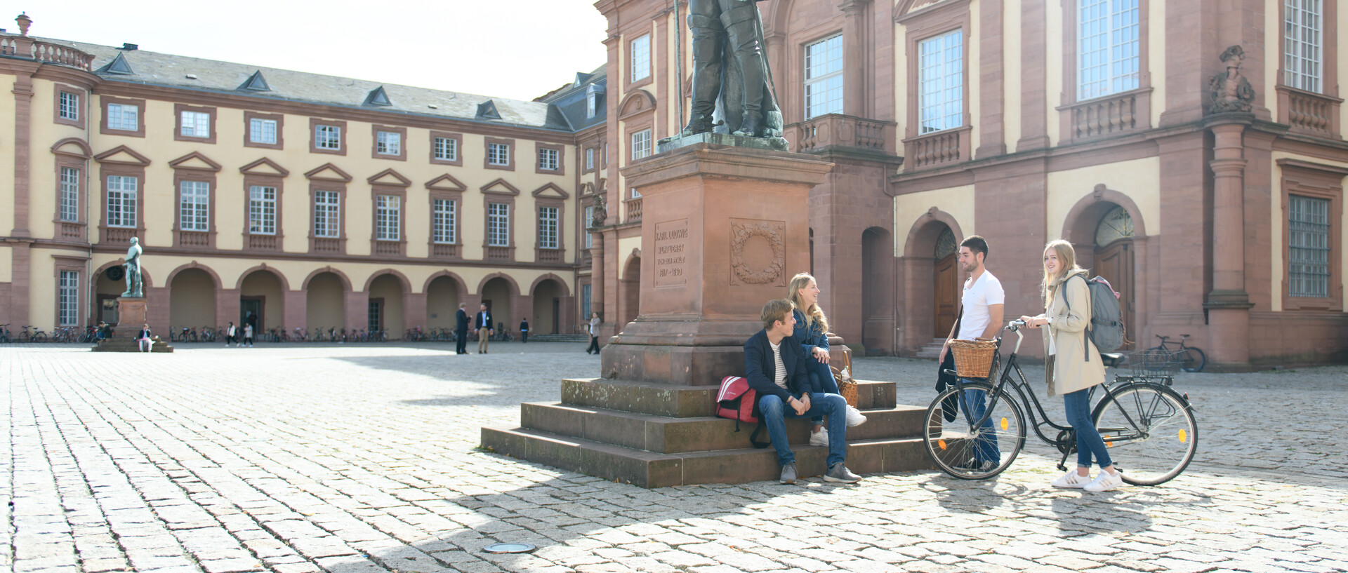 Eine Gruppe Studierender an der Statue auf dem Ehrenhof, zwei Studierende sitzen auf den Stufen, zwei stehen davor. Eine Studierende schiebt ein Fahrrad.