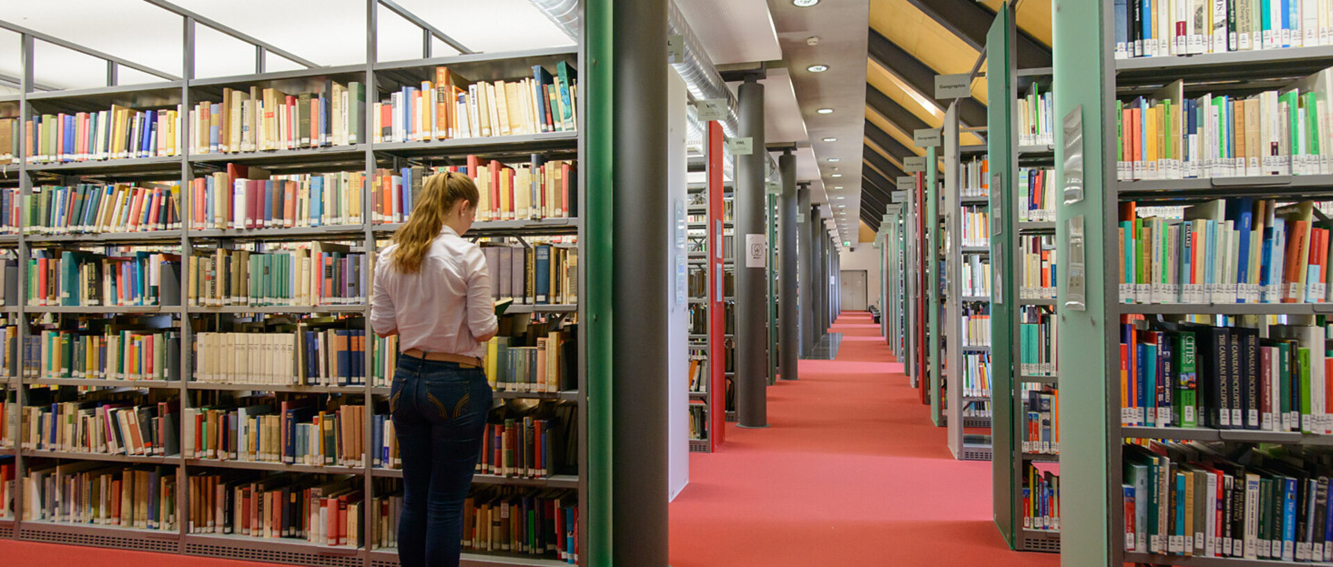 Eine Studentin steht vor einem Bücherregal im Bibliotheksbereich Schloss Ehrenhof. Daneben und dahinter sind weitere Bücherregale mit Regalblenden in Rot- und Grüntönen. 