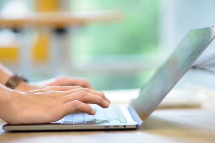 Eine Person, deren Hände im Bild sind, schreibt am Laptop. Das Laptop steht auf einem Tisch, im Hintergrund ist ein Stehtisch.