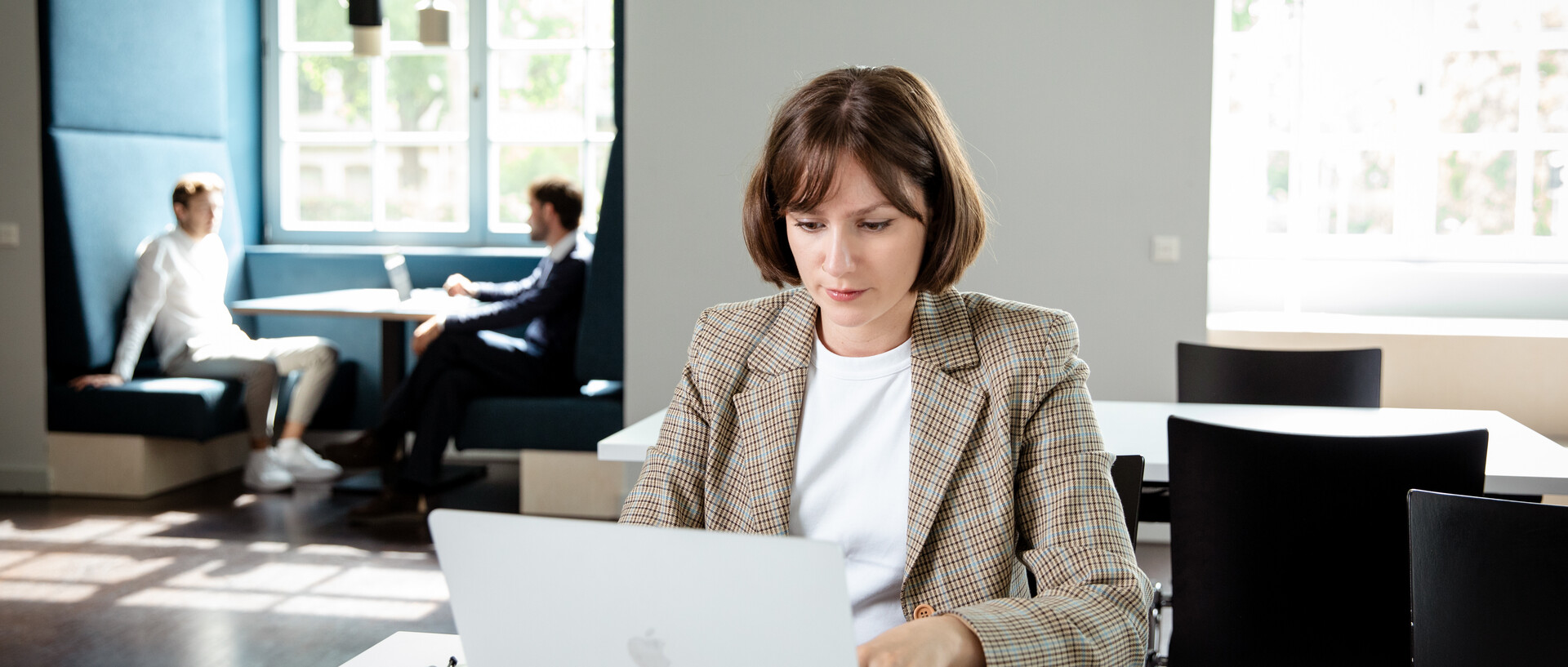Eine Frau sitzt an einem Tisch im ExpLAB im Bibliotheksbereich Schloss Schneckenhof. Sie schreibt an einem Laptop. Im Hintergrund sitzen zwei Personen in einer Fensternische.