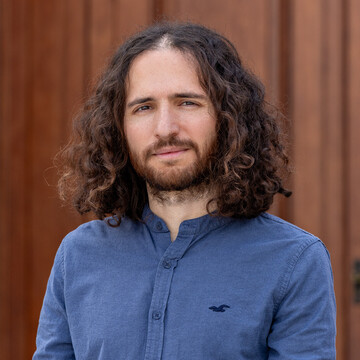 Christos Sidiropoulos hat lockige lange Haare und einen Bart. Er trägt ein blaues Hemd.