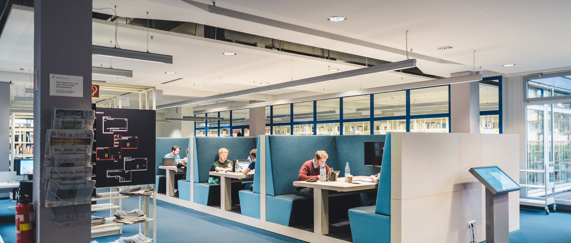 Studierende lernen in den blauen Kuben im Bibliotheksbereich A5. Links im Bild sind Zeitungen und ein Bücherregal.