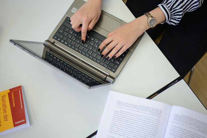 Eine Person, deren Hände im Bild sind, schreibt an einem Laptop. Neben dem Laptop liegen Bücher auf dem Tisch. Die Person trägt eine Uhr am rechten Handgelenk und hat schwarz lackierte Fingernägel.