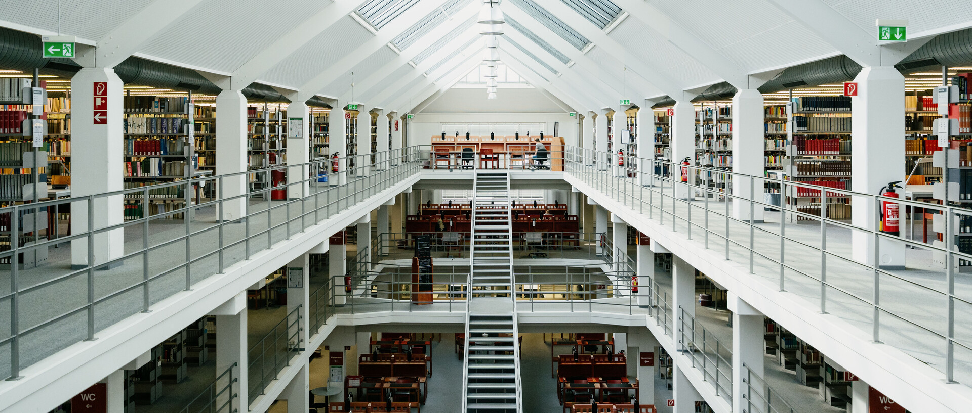 Bibliotheksbereich A3 mit Arbeitsplätzen im Erdgeschoss, dem ersten und zweiten Stock mit Bücherregalreihen und langer Treppe vom Erdgeschoss in den zweiten Stock mit sonnigem Glasdach.