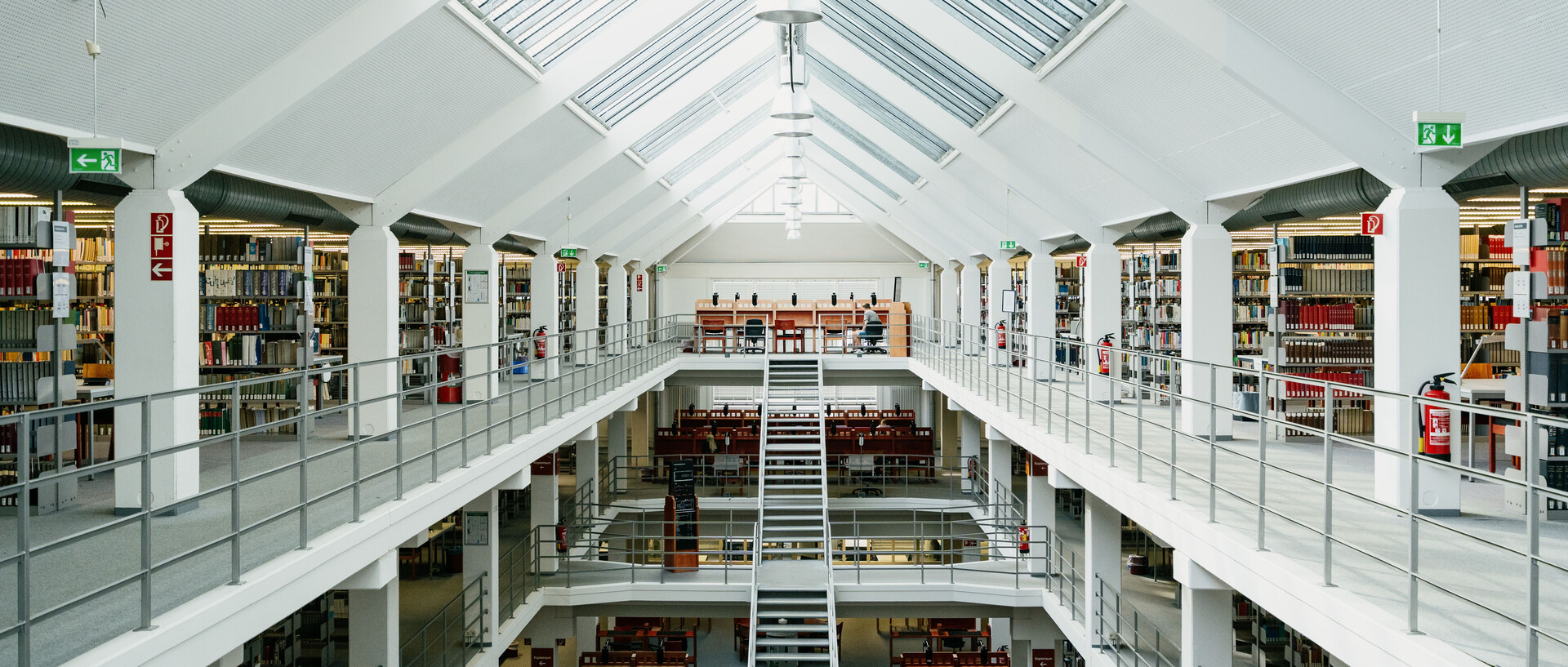 Bibliotheksbereich A3 mit Arbeitsplätzen im Erdgeschoss, dem ersten und zweiten Stock mit Bücherregalreihen und langer Treppe vom Erdgeschoss in den zweiten Stock mit sonnigem Glasdach.