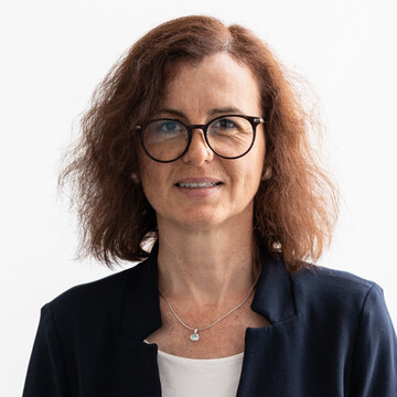 Isabelle Otto hat halblanges braunes Haar, sie trägt eine Brille mit schwarzem Gestell, ein weißes Oberteil, ein dunkelblaues Jacket und eine silberne Halskette mit hellblauem Anhänger.