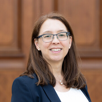 Angela Leichtweiß hat langes braunes Haar, sie trägt eine Brille, Perlenohrringe, eine Kette, ein weßes Oberteil und ein dunkles Jackett. 