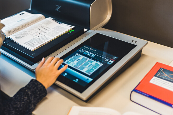 Eine Studentin benutzt einen Scanner in der Bibliothek. Ein Buch liegt auf der Scanfläche und sie nimmt die Einstellungen am Bedienfeld vor. Ein weiteres Buch liegt rechts neben dem Scanner auf dem Tisch. 