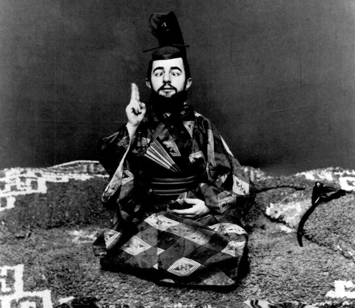 Ausschnitt einer Fotografie von Maurice Guibert aus dem Jahr 1892. Zu sehen ist der Maler Henri de Toulouse-Lautrec. Er trägt japanische Tracht, sitzt auf einem Teppich, hat zwei Finger der rechten Hand erhoben und schielt. Das Schielen ist eine stereotype, rassistische Darstellung asiatischer Personen. 