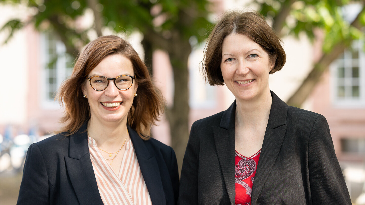 Sabine Gehrlein und Annette Klein stehen nebeneinander vor dem Schloss. Sabine Gehrlein hat schulterlanges rotbraunes Haar, sie trägt eine Brille, Perlenohrringe, eine Kette, ein rosé-weiß gestreiftes Hemd und ein dunkles Jackett. Annette Klein hat halblanges braunes Haar, sie trägt ein rotes Oberteil mit floralem Muster und ein schwarzes Jackett.