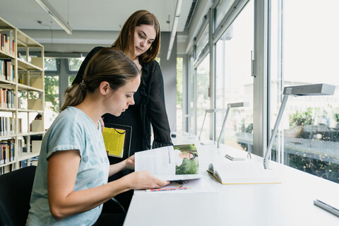 Zwei junge Frauen sehen sich gemeinsam an einem Tisch am Fenster im Bibliotheksbereich A5 eine Zeitschrift an.