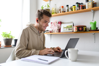 Student in hellbraunen Kapuzenpulli sitzt in seiner Wohnung vor dem Laptop und hat Kopfhörer an. Auf dem Tisch steht eine Tasse und liegt ein Notizblock. Link: Promotion