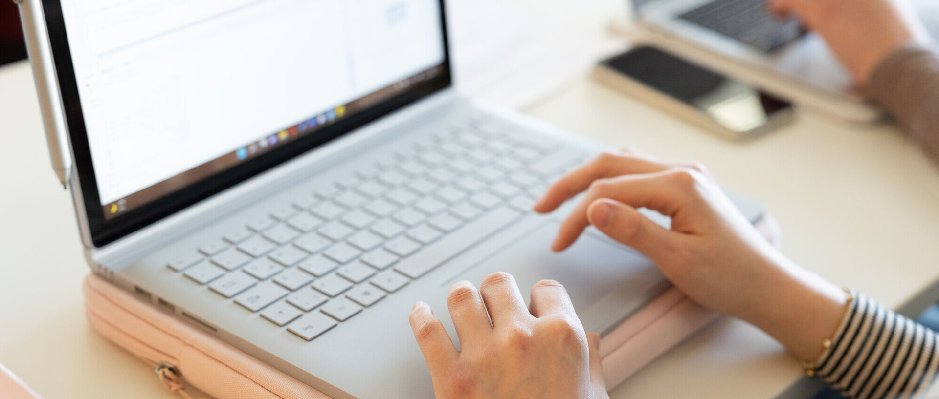 Ein Person, deren Hände im Bild sind, schreibt an einem Laptop. Das Laptop steht auf einer hellrosa Hülle auf einem Tisch.